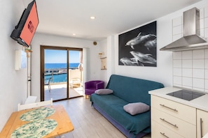 Una sala moderna con un sofá color turquesa, una mesa de comedor con mantel floral, una cocina con campana extractora de acero inoxidable y un ventanal con vistas a una masa de agua.
