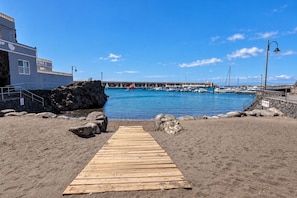 Un sentier en bois mène à une plage rocheuse, avec un ciel bleu dégagé et une marina en arrière-plan.