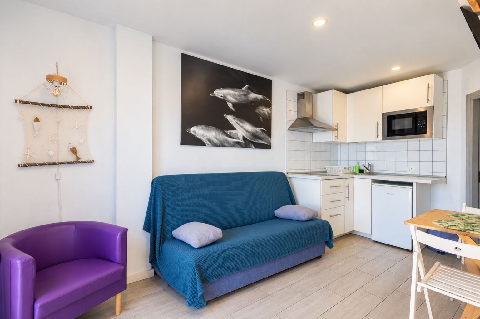 A compact living space with a sofa, a purple chair, a dining table, and a kitchen area with white cabinets and a microwave.
