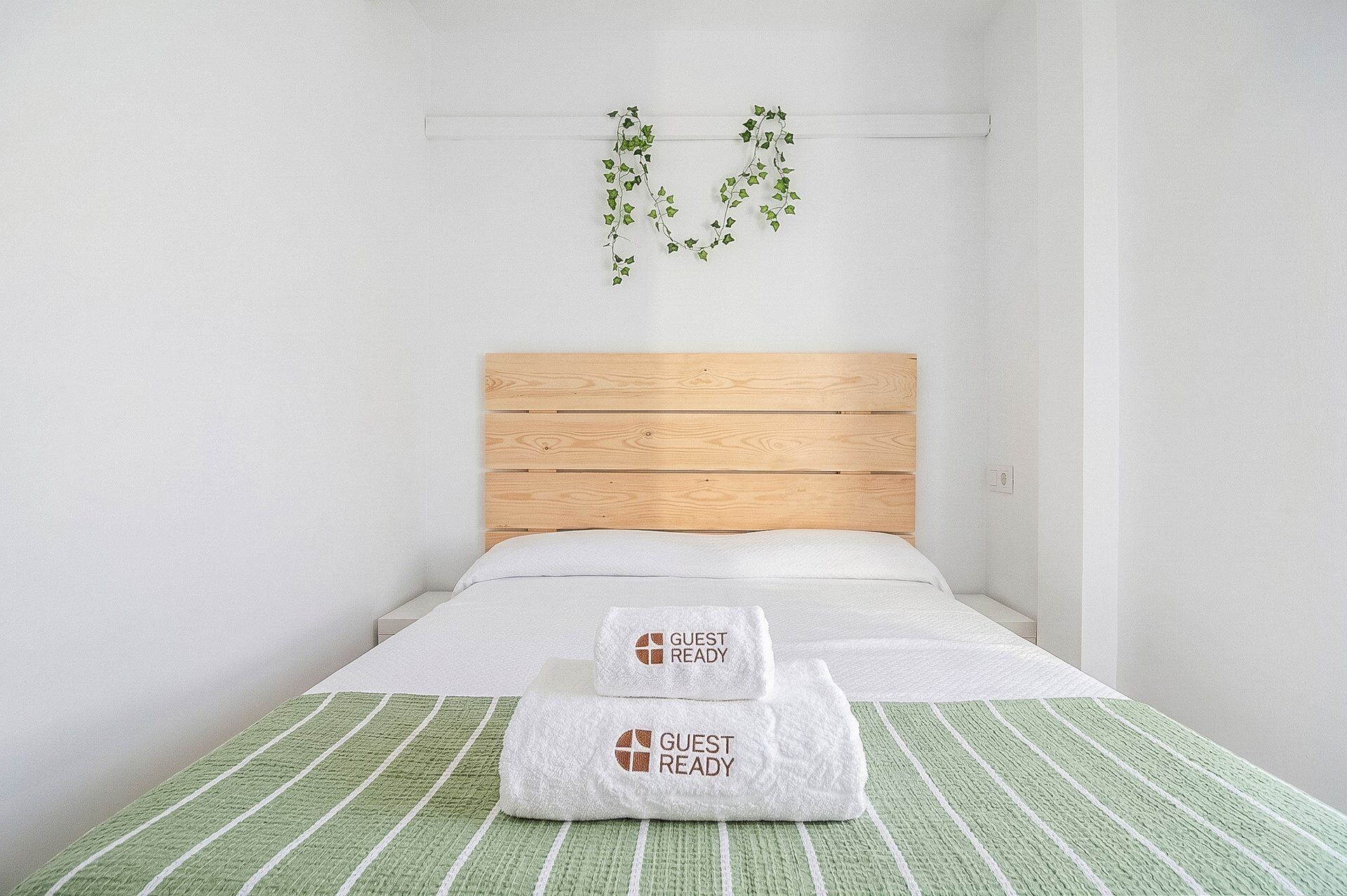 Ein Einzelbett mit Holz Kopfteil, zwei weiße Handtücher mit dem Aufdruck 'GUEST READY' und eine grün gestreifte Bettdecke.