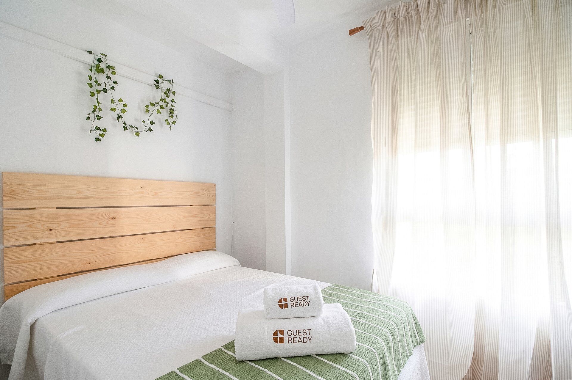 Una cama bien hecha con cabecera de madera, manta a rayas verde y blanca, y dos toallas blancas con el texto 'GUEST READY'.