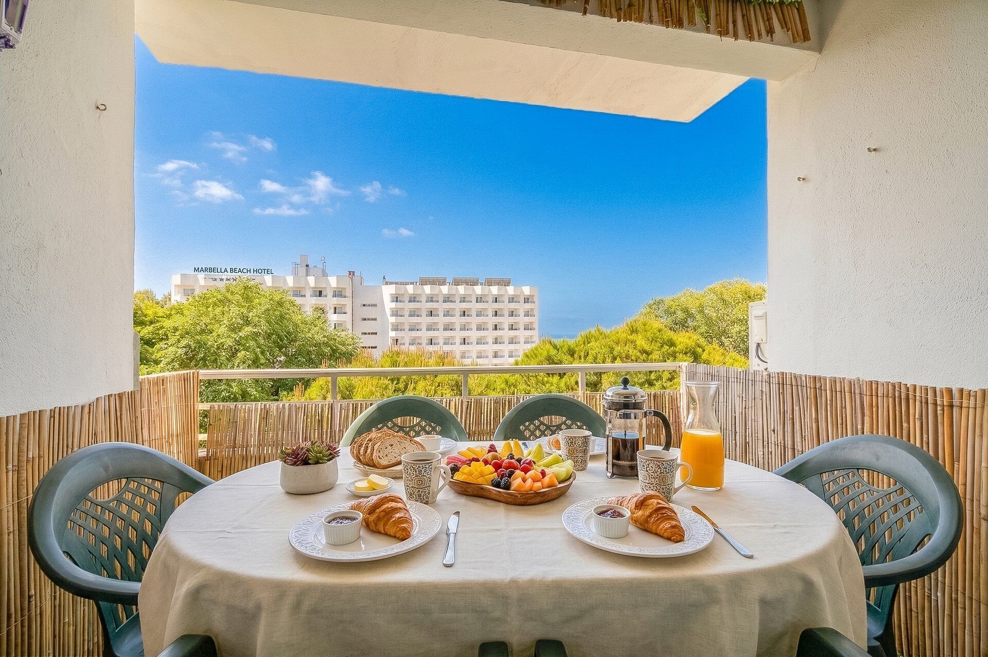 Een balkon met een tafel gedekt voor het ontbijt, met daaronder croissants, fruit en een drankje, met uitzicht op een stadsgezicht met gebouwen en groen.