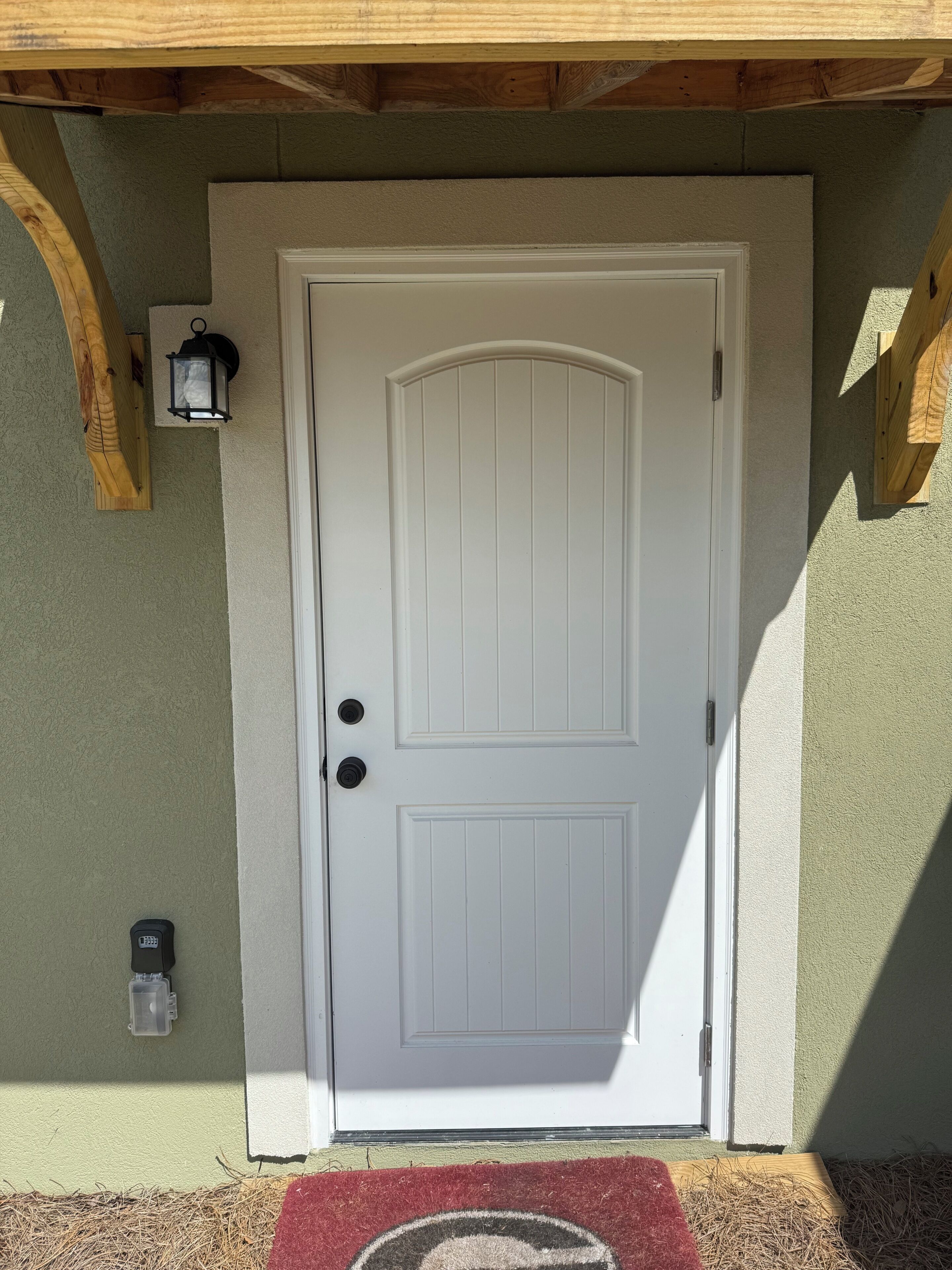 A white door with a keyhole and a doorbell, a mailbox, and a red doormat with a circular design.