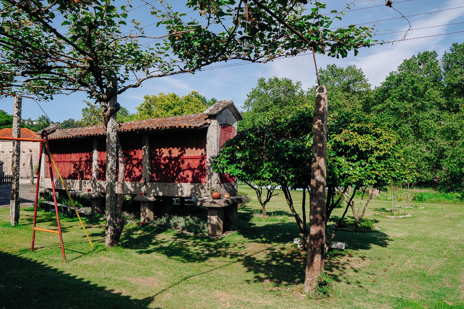 Un bâtiment traditionnel en pierre, doté d’une façade rouge et soutenu par des piliers de pierre, situé dans un espace verdoyant, parsemé d’arbres, sous un ciel dégagé.