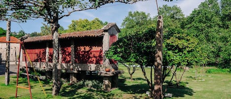A traditional stone building with a red facade, supported by stone pillars, in a grassy area with trees and a clear sky.