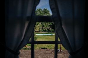 A view through a window with curtains, featuring a swimming pool surrounded by grass and trees. - Casa de Requeixo - 4-bedroom vacation home with a shared pool (Povoa De Lanhoso)