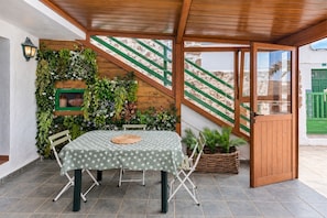 Een terras buiten met een tafel, stoelen, een groen geruit tafelkleed, een rieten mand en een houten pergola.