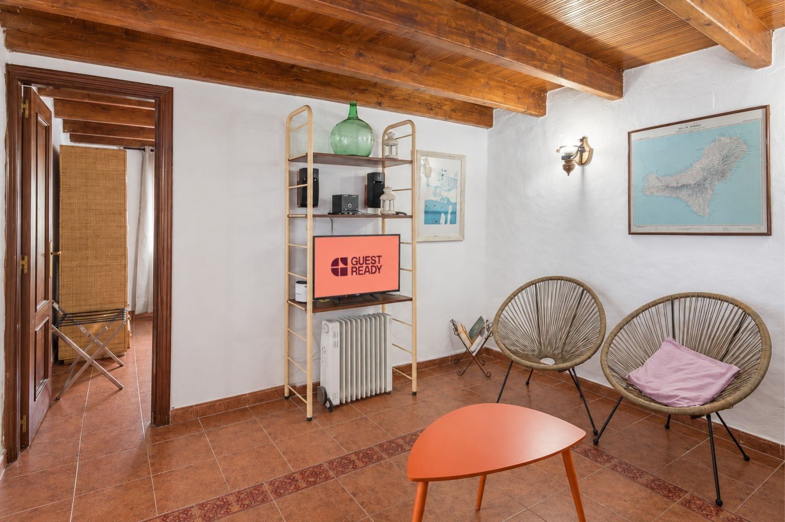 A room with a wooden ceiling, a radiator, a shelf with a 'GUEST READY' sign, two wicker chairs, and a framed map on the wall.