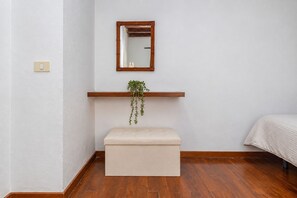 Een kamer met een houten vloer, een witte muur, een houten plank met een hangende plant, een klein beigekleurd bankje en een bed met een witte deken.