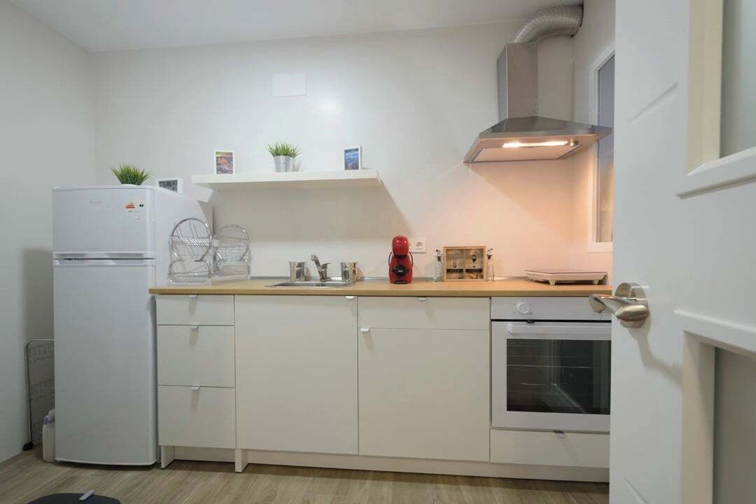 A compact kitchen with white cabinets, a built-in oven, a refrigerator, and a countertop with a kettle and a red coffee maker.