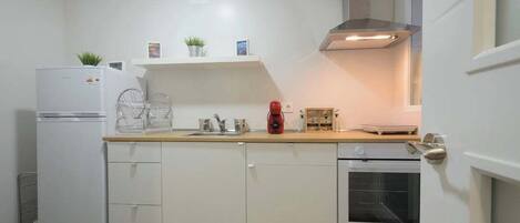 A compact kitchen with white cabinets, a built-in oven, a refrigerator, and a countertop with a kettle and a red coffee maker.