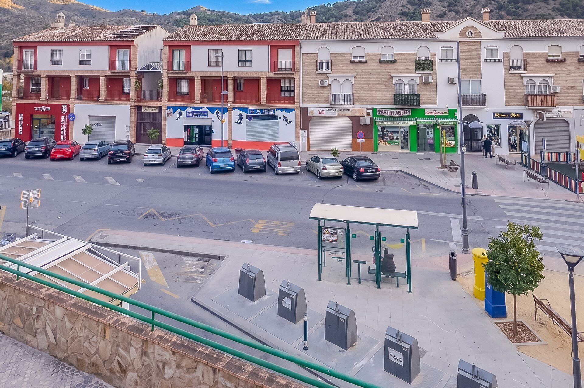 A street view with parked cars, a bus stop, and buildings with commercial establishments.