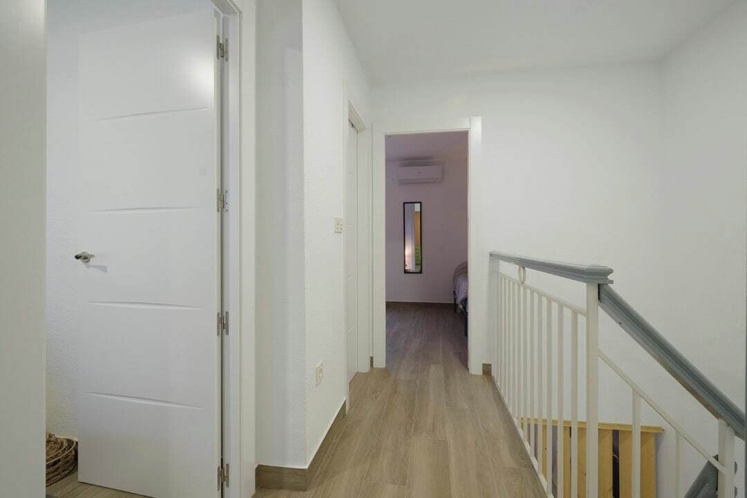 Un pasillo con suelo de madera, paredes blancas y una puerta blanca a la izquierda.