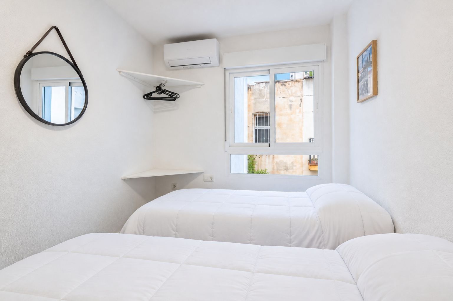 A bedroom with two beds, a hanging mirror, an air conditioning unit, a window with a view of buildings, and a picture frame on the wall.