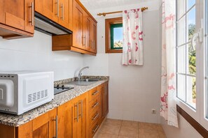 A compact kitchen with wooden cabinets, a white microwave, a sink, and a window with a floral curtain. - GuestReady - Colon I 11 2 pax (Los Quemados)
