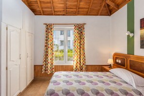 Une chambre avec un plafond en bois, un lit recouvert d’une couvre-lit à pois, une table de chevet avec une lampe et une fenêtre ornée de rideaux à carreaux.