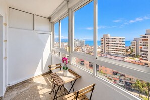 A balcony with a table and chairs, a vase with flowers, and a view of a cityscape with buildings and the sea. - GuestReady - Las Dalias Torremolinos Comfort (Torremolinos)
