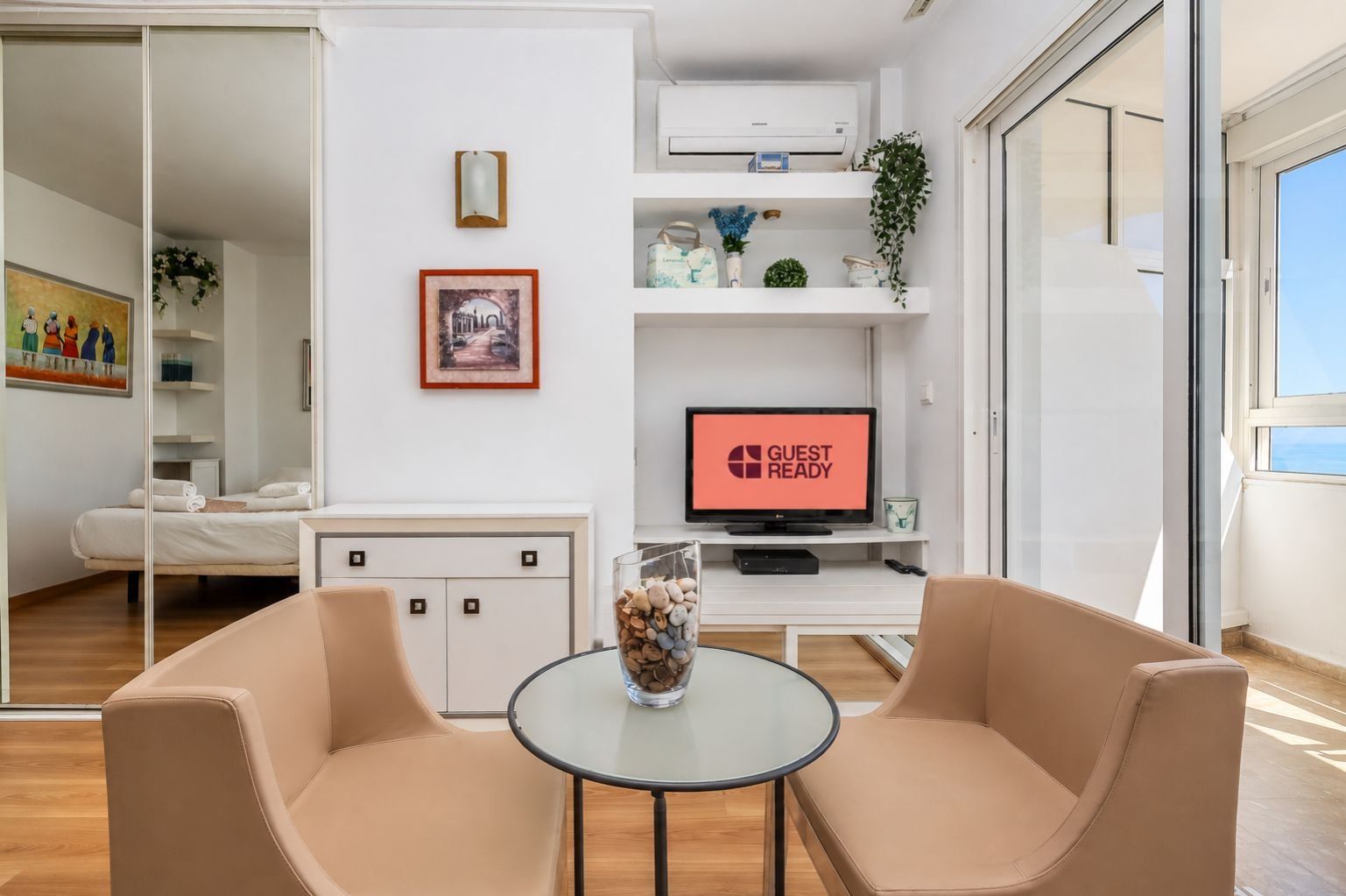 Una camera d'albergo moderna con un letto, un tavolino in vetro con sedie, una TV che mostra 'GUEST READY' e una porta scorrevole in vetro che dà all'esterno.