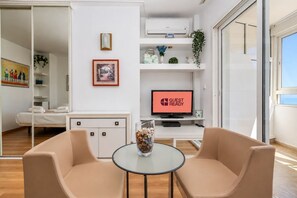 A modern hotel room with a bed, a glass table with chairs, a TV displaying 'GUEST READY', and a sliding glass door to the outside. - GuestReady - Las Dalias Torremolinos Comfort (Torremolinos)