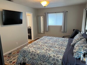 Ein Schlafzimmer mit einem Bett, einem an der Wand montierten Fernseher, einem Fenster mit Vorhängen und einer Tür zu einem Badezimmer.