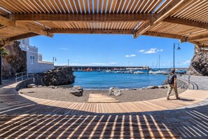 A wooden deck with a view of a harbor, a rocky shoreline, and a building in the background. - GuestReady - Beachfront Apartment Marina Views (La Restinga)