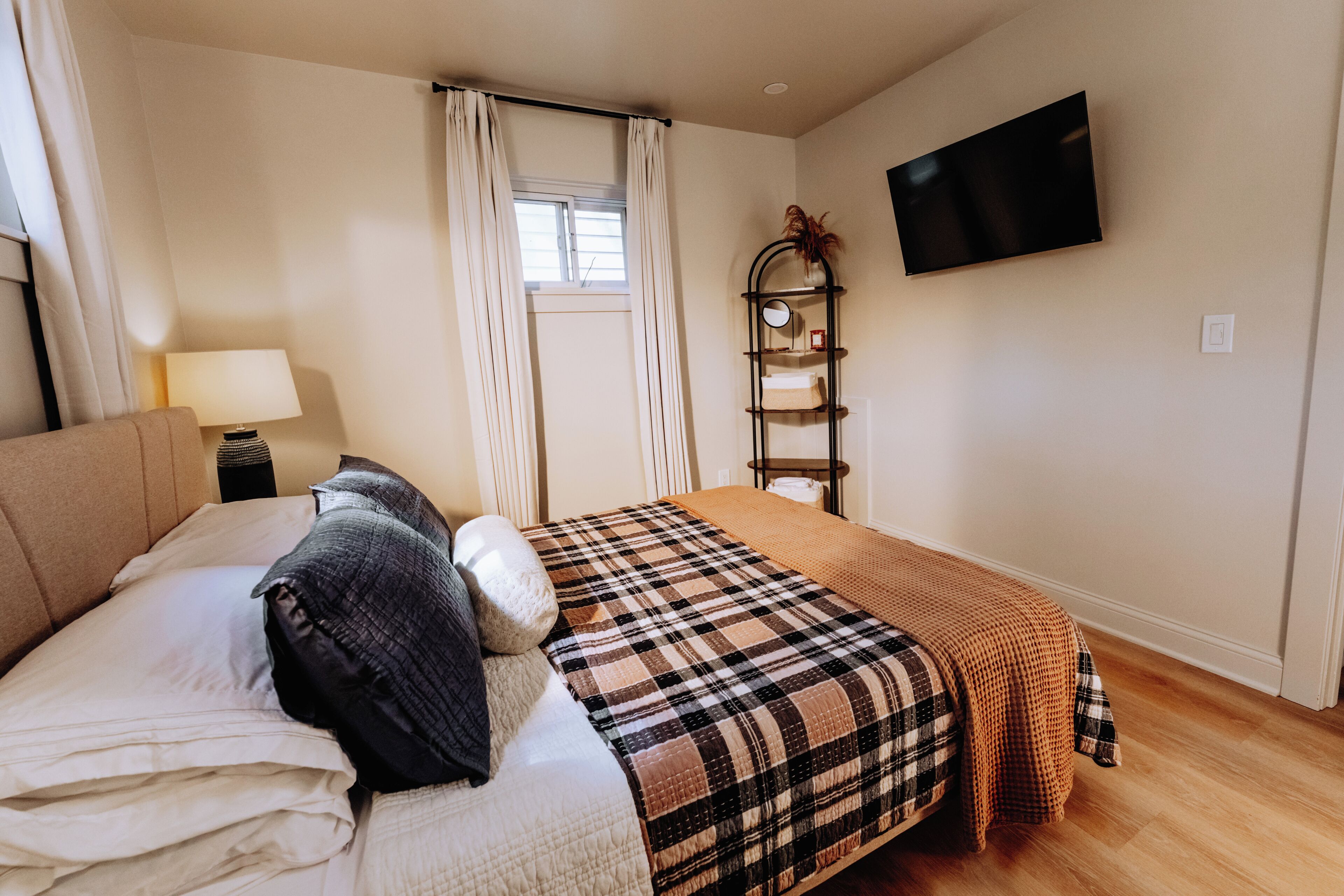 Une chambre à coucher avec un lit, une table de chevet avec une lampe, une fenêtre avec des rideaux, un téléviseur fixé au mur et une étagère avec des objets décoratifs.