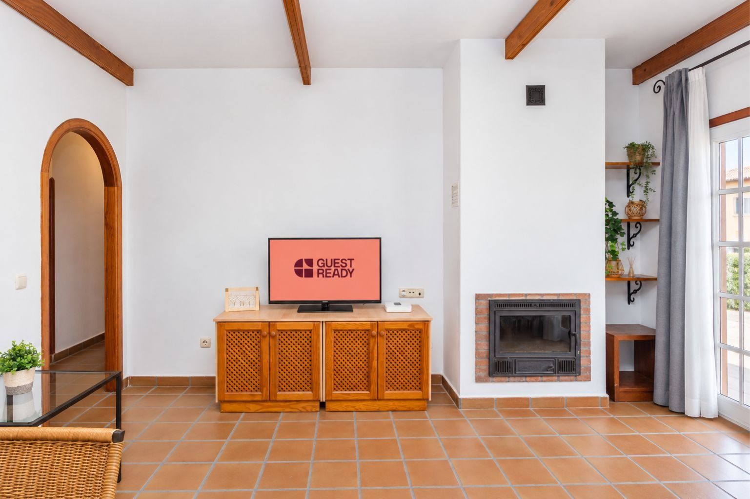 A living room with a television displaying 'GUEST READY,' a fireplace, a wooden cabinet, and a large window with curtains.