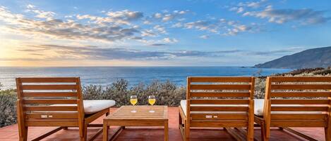 A terrace with two wooden chairs and a table, two glasses of wine, and a view of the sea and hills.