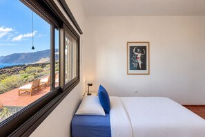 A bedroom with a bed, a window with a view of the sea and mountains, a framed picture on the wall, and a small table with chairs outside the window. - GuestReady - Beachfront Bungalow Sunsets (Los Llanillos)