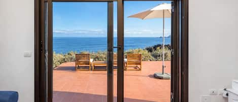 Une pièce dotée d’une porte-fenêtre coulissante donnant sur une terrasse extérieure avec vue sur la mer, un banc et un parasol.