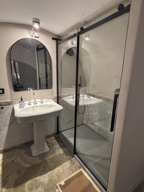 A bathroom with a pedestal sink, a glass-enclosed shower, and a mirror with a curved arch. - Vaulted ceilings just renovated loft apartment Tarquinia old town (Tarquinia)