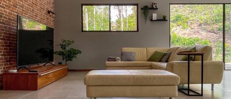 Una sala de estar con pared de ladrillo, televisor, sofá, mesa de centro y ventana con vista a zonas verdes.