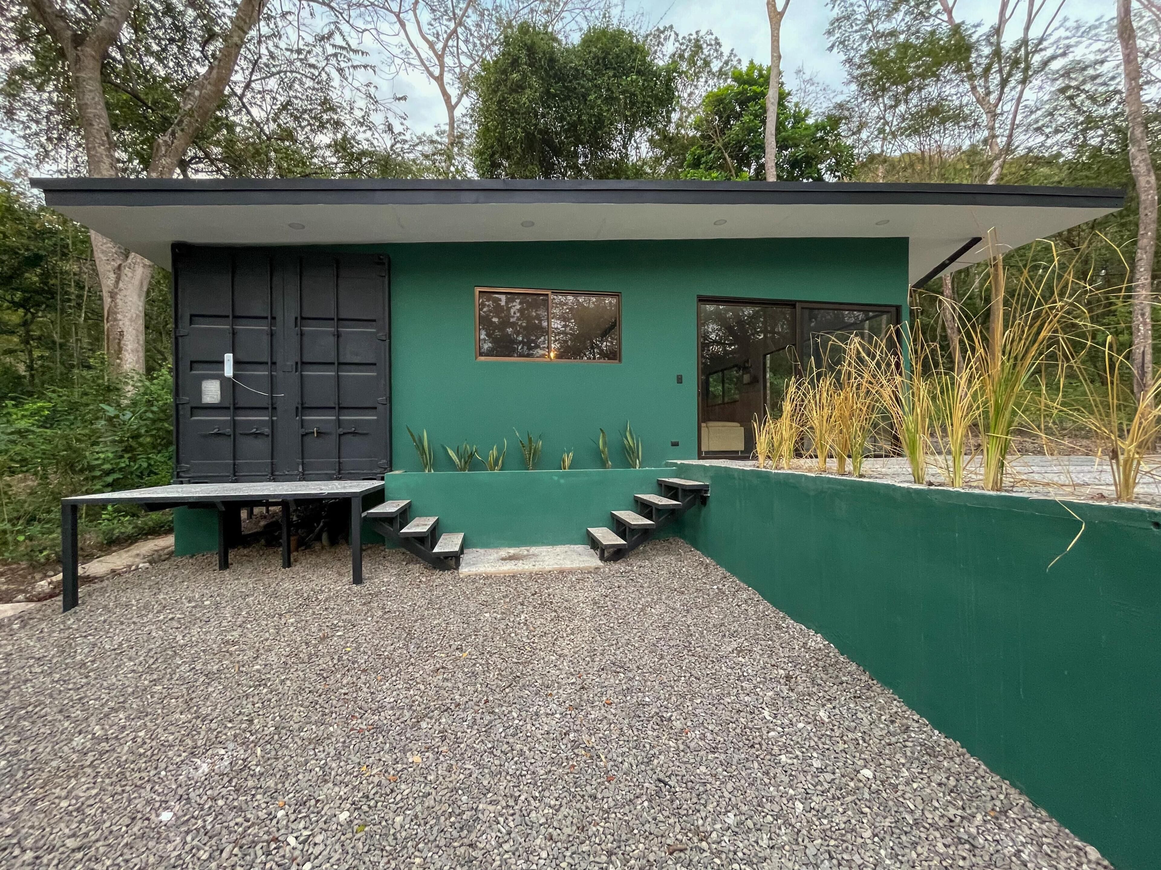 Un edificio moderno con facciata verde, una porta nera e una piccola finestra. Un'area con ghiaia, una panchina e gradini che conducono all'ingresso.