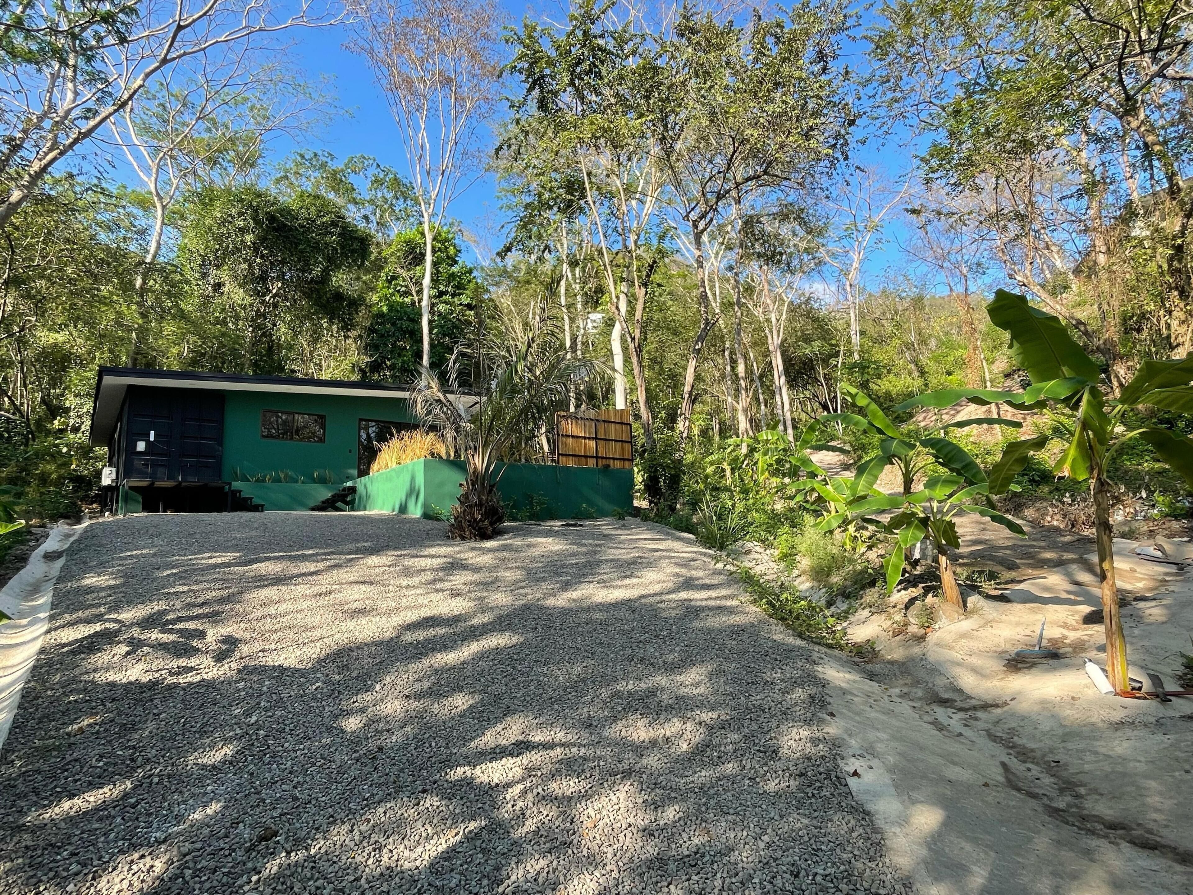 Un camino de grava conduce a un edificio verde con garaje, rodeado de árboles y vegetación.