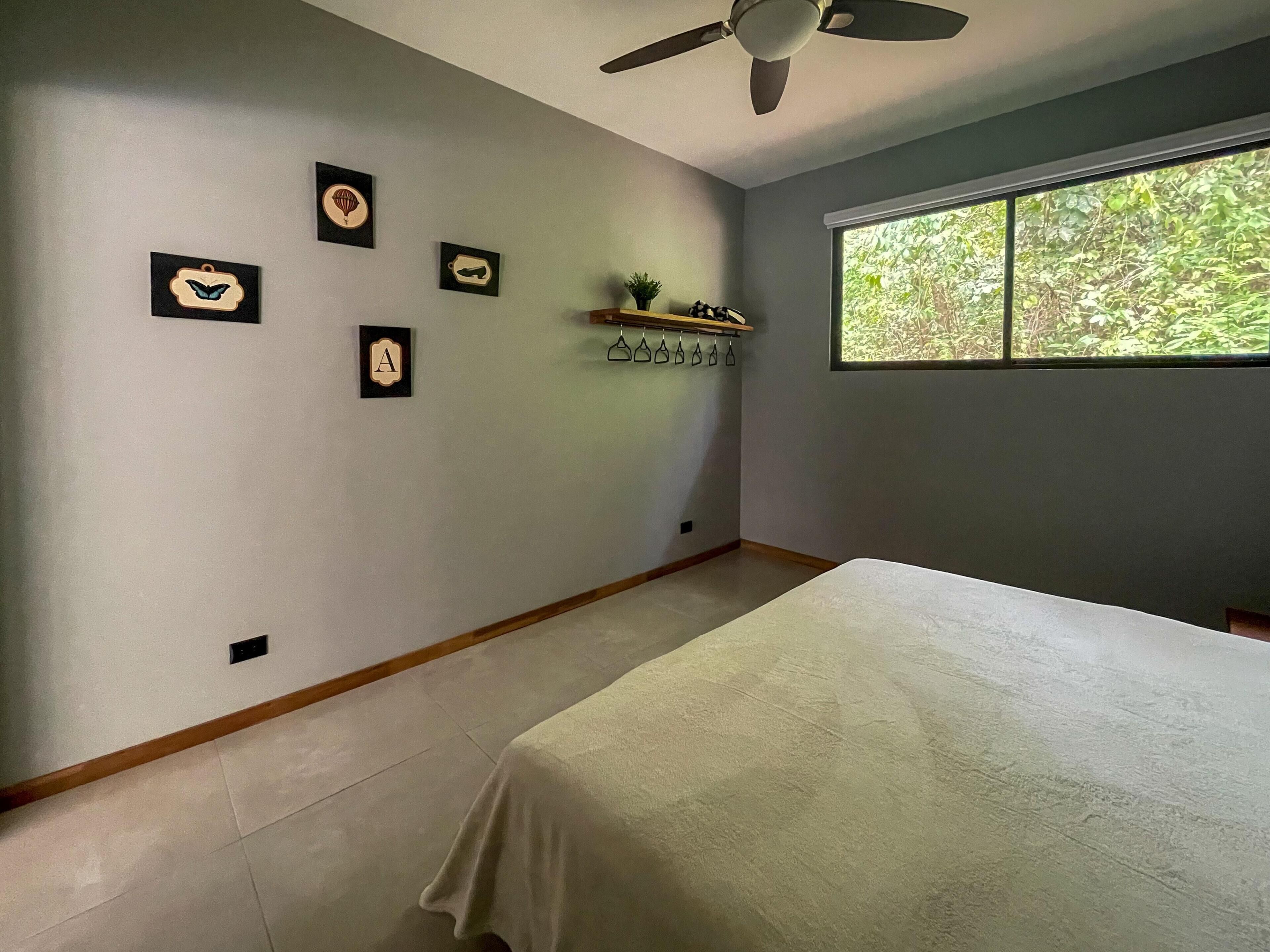 Une chambre à coucher avec un lit, un ventilateur de plafond, une fenêtre donnant sur un paysage verdoyant et des œuvres d’art accrochées au mur.