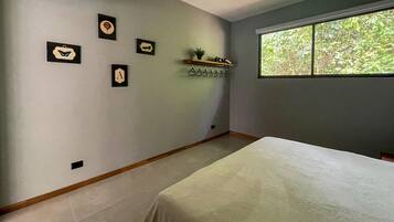 Une chambre à coucher avec un lit, un ventilateur de plafond, une fenêtre donnant sur un paysage verdoyant et des œuvres d’art accrochées au mur.