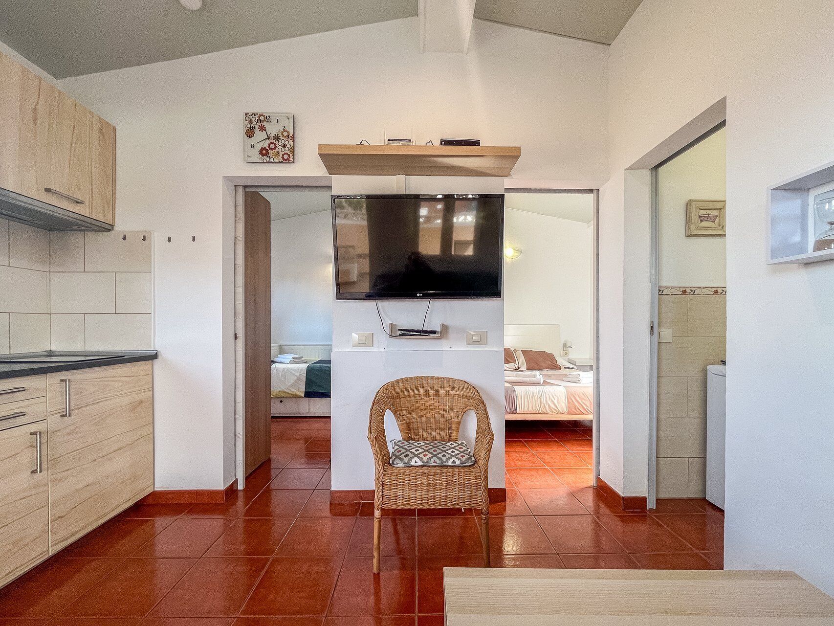 A compact living space with a kitchenette, a television mounted above a central table, and a wicker chair in the foreground.
