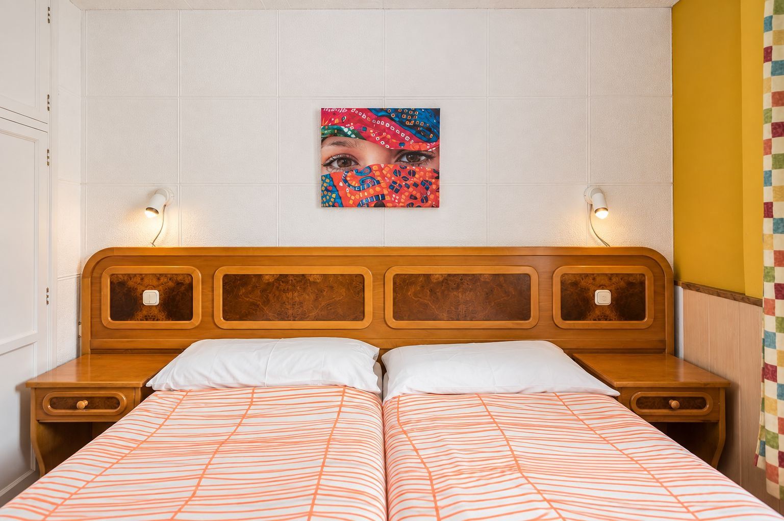 Una habitación de hotel con cabecera de madera, dos mesitas de noche, una cama con ropa de cama a rayas, un cuadro colgado en la pared y una lámpara a cada lado de la cama.