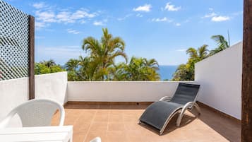 Una terraza con una mesa blanca y dos sillas, una silla de descanso negra, y vistas a palmeras y al océano.