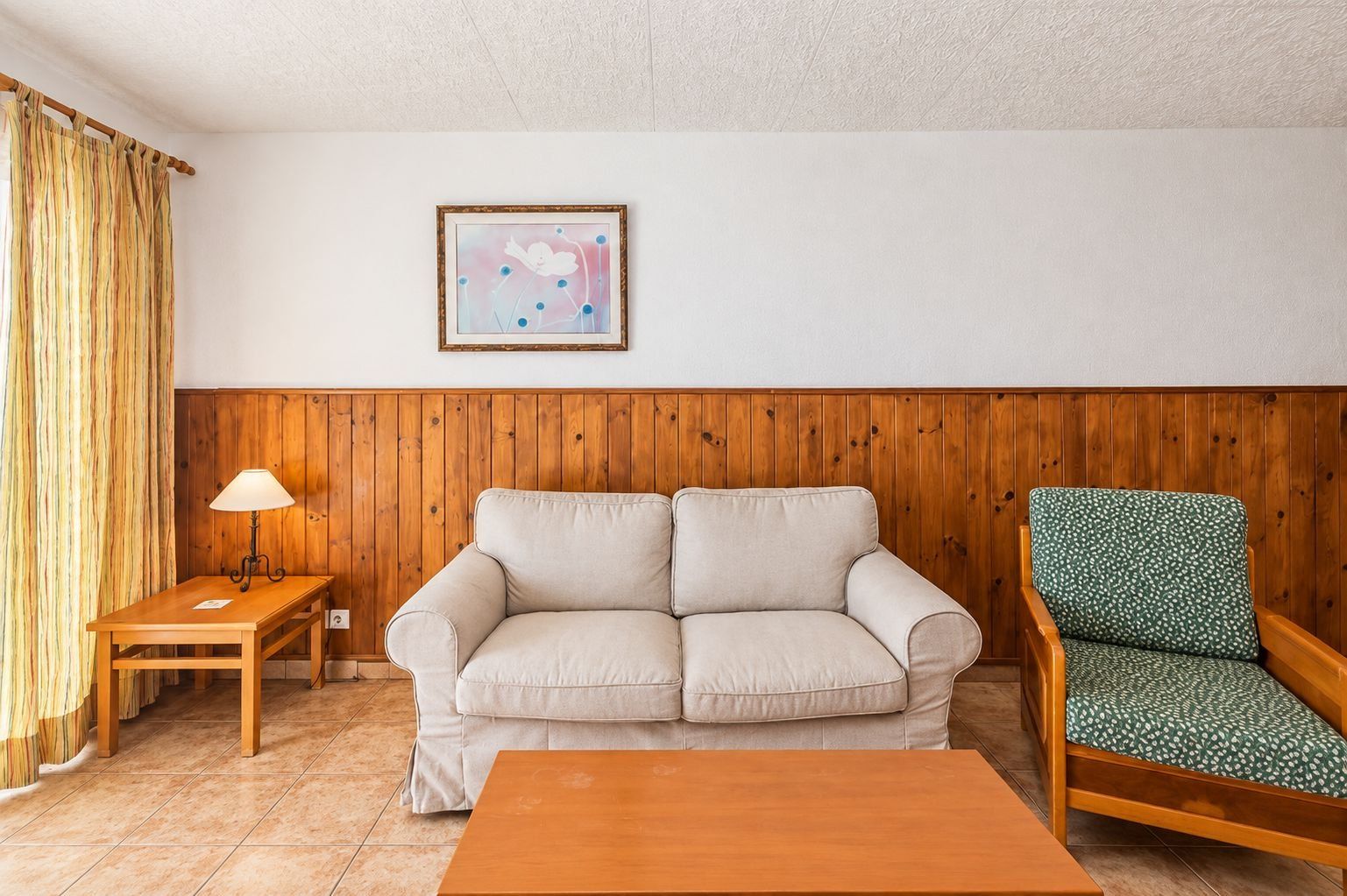 Una sala de estar con un revestimiento de madera, un sofá color beige, un sillón estampado en verde, una mesa de centro de madera y una lámpara sobre una mesita auxiliar.