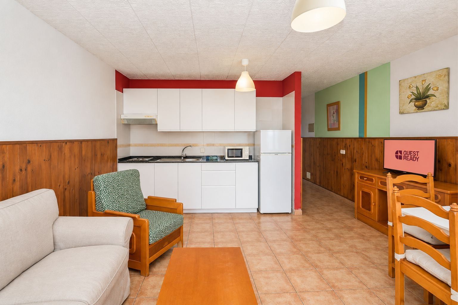 A compact living space with a kitchenette, a sofa, a chair, a small table, and a television displaying 'GUEST READY'.
