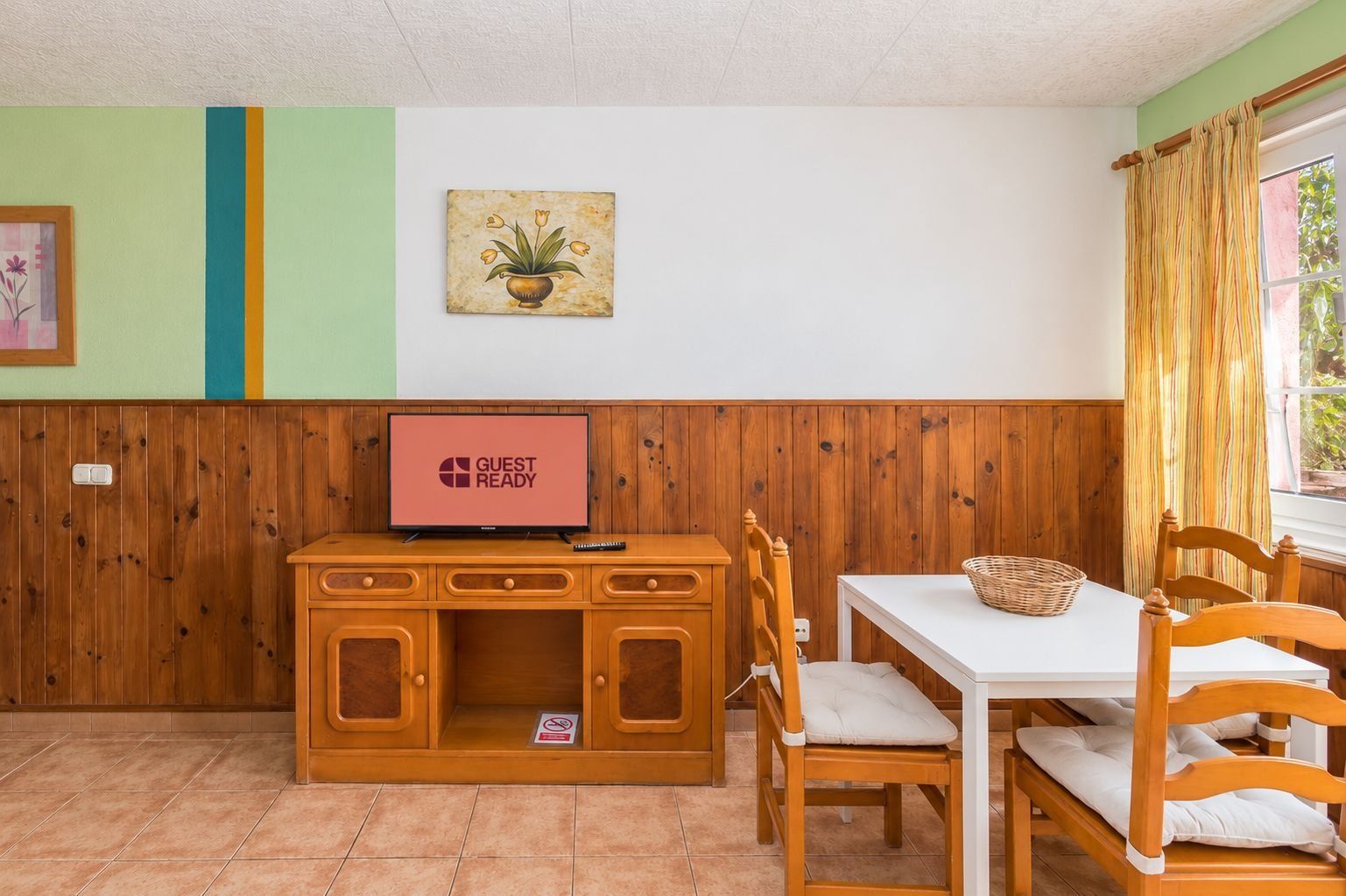 Una habitación con un armario de madera, un televisor mostrando 'GUEST READY', una mesa de comedor con sillas y una ventana con cortinas.