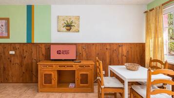 Una habitación con un armario de madera, un televisor mostrando 'GUEST READY', una mesa de comedor con sillas y una ventana con cortinas.