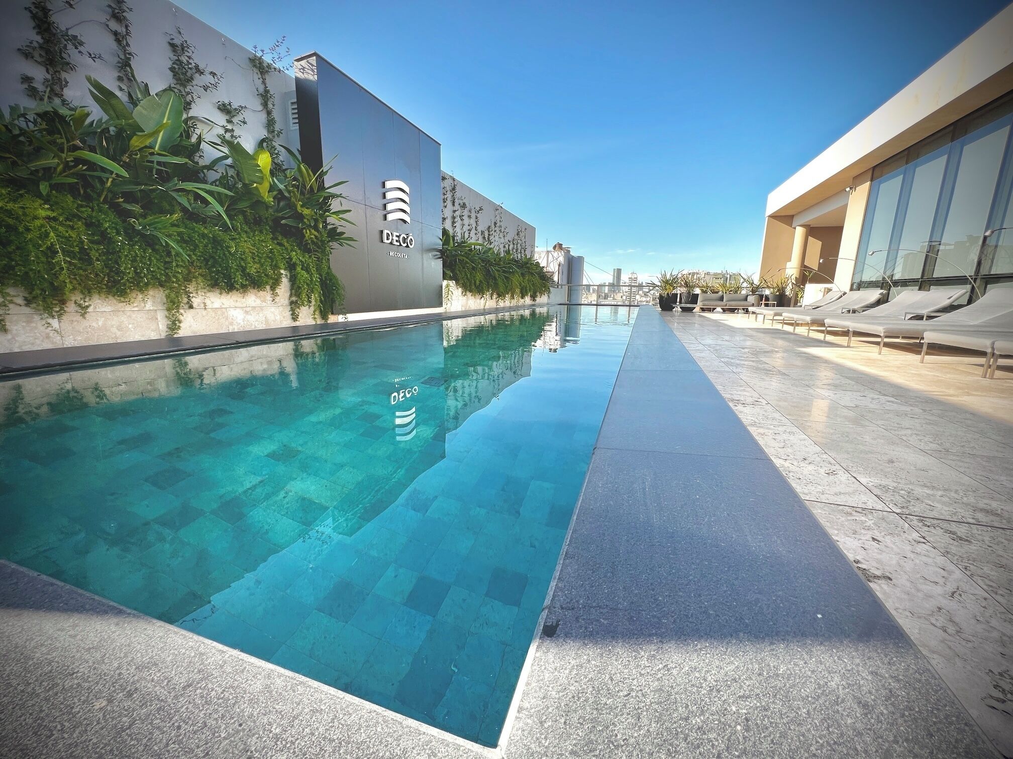 Une piscine à l’eau d’un bleu cristallin, entourée d’un bâtiment moderne doté d’une façade en verre et d’un espace extérieur aménagé pour se détendre.