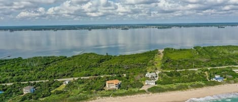 Luftaufnahme einer Küstenlandschaft mit Strand, weiter Wasserfläche und dichtem Grün mit Gebäuden.