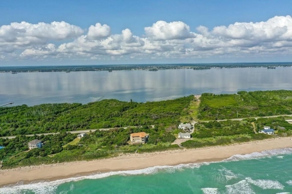 An aerial view of a coastal area with a beach, a large body of water, and a dense green landscape with buildings.
