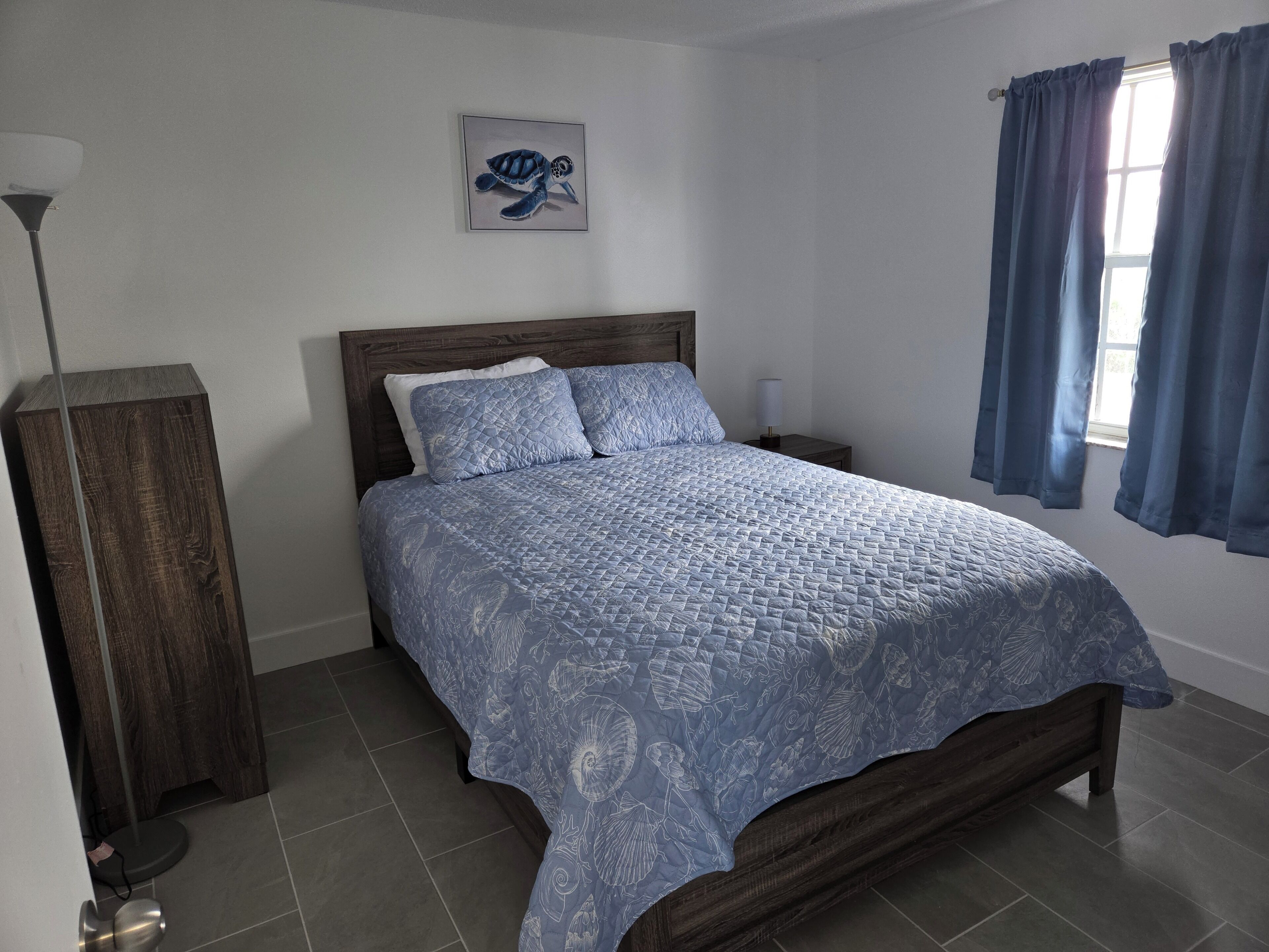 A bedroom with a bed, a nightstand, a lamp, a picture on the wall, and a window with curtains.