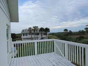 A white wooden deck with a white railing, overlooking a house with a balcony and palm trees, with a beach and ocean in the background.
