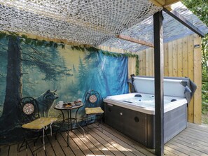 A hot tub in a covered outdoor area with a forest mural backdrop, a table with chairs, and a wooden deck. - Squirrels Lodge (South Molton)