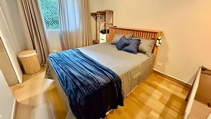 Un dormitorio con cama, cabecera de madera, mesita de noche pequeña, una silla y una ventana con cortinas.